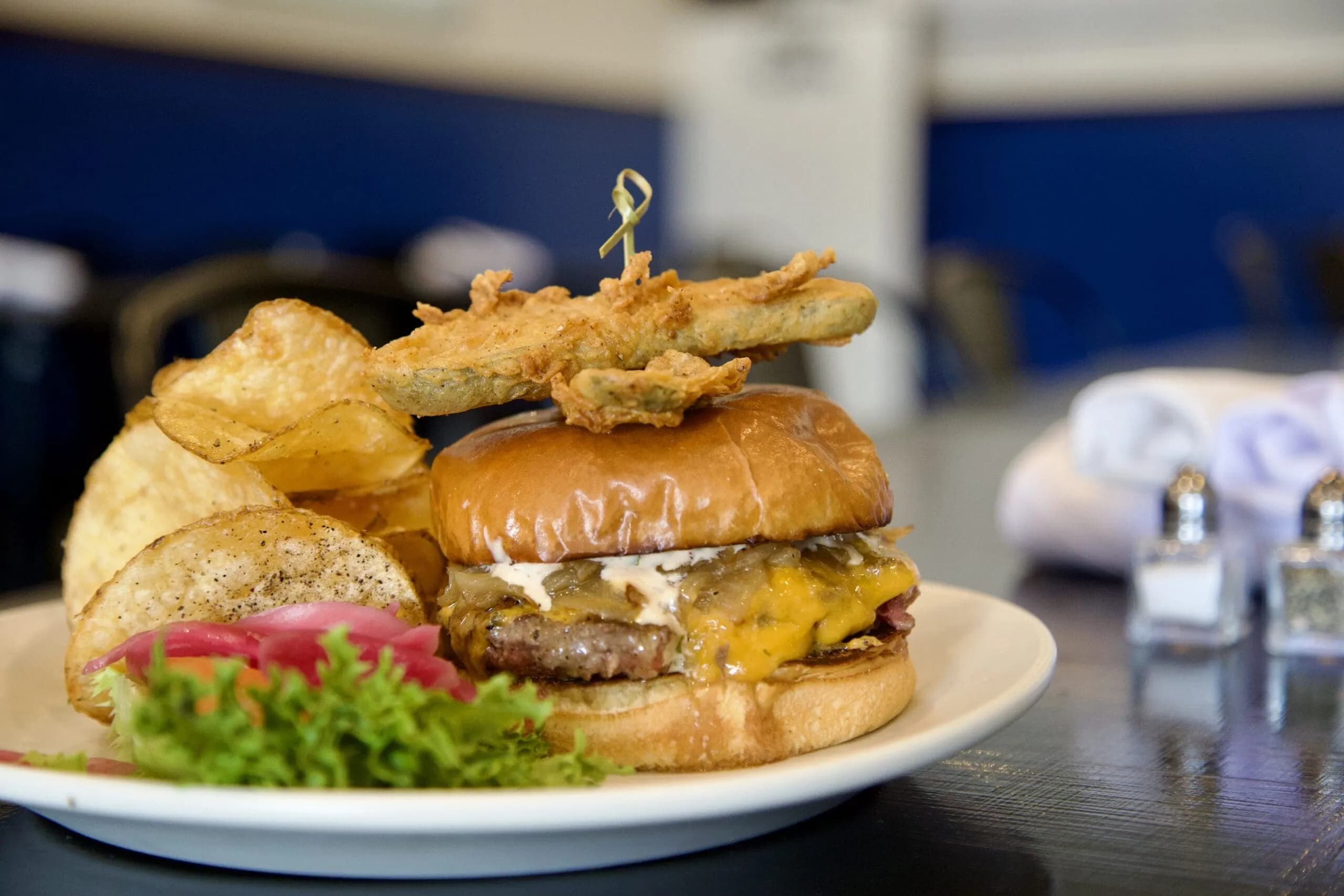 A stacked cheeseburger topped with fried pickles and served with kettle chips — one of the hearty dishes spotlighted in Tucson Foodie’s Passport for local restaurants.