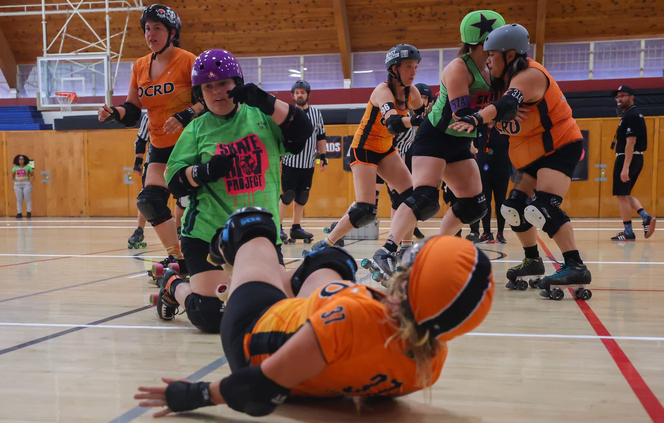 An Orange County Roller Derby skater in orange tumbles to the track as members of Skate Riot Project (green) and OCRD contest a jam at Broadway Recreation Center in Mesa on June 27, 2026.