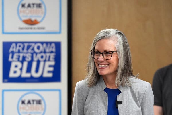 A female politician smiles during a campaign event with “Arizona Let’s Vote Blue” signage behind her, shown alongside reporting that a potential bipartisan running mate may join the ticket.