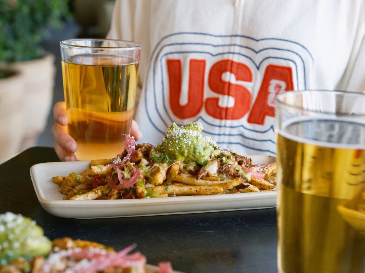 A U.S. soccer fan in a 'USA' shirt with Blanco Cocina + Cantina’s loaded fries and a pint — illustrating the $12 fries-and-beer World Cup special called out in the article.