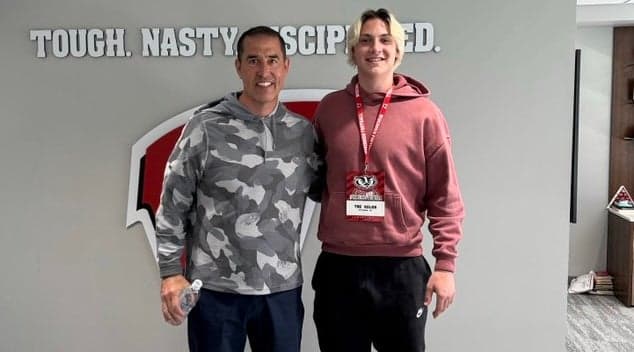 A visitor poses with a Wisconsin staff member in front of the Badgers' 'TOUGH. NASTY. DISCIPLINED.' wall inside the program's facility during an unofficial visit to Madison.