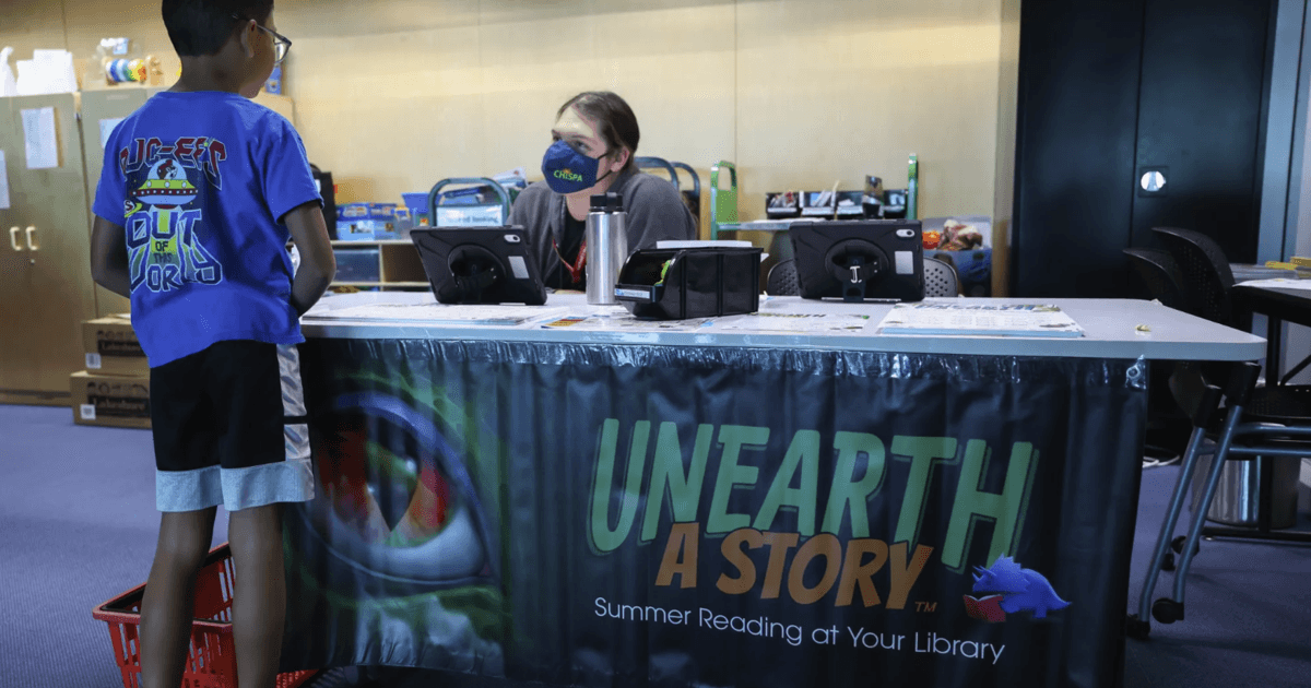 Library staff register a child at the 'Unearth a Story' summer reading table — Phoenix libraries' dinosaur-themed effort to combat the summer reading slump.