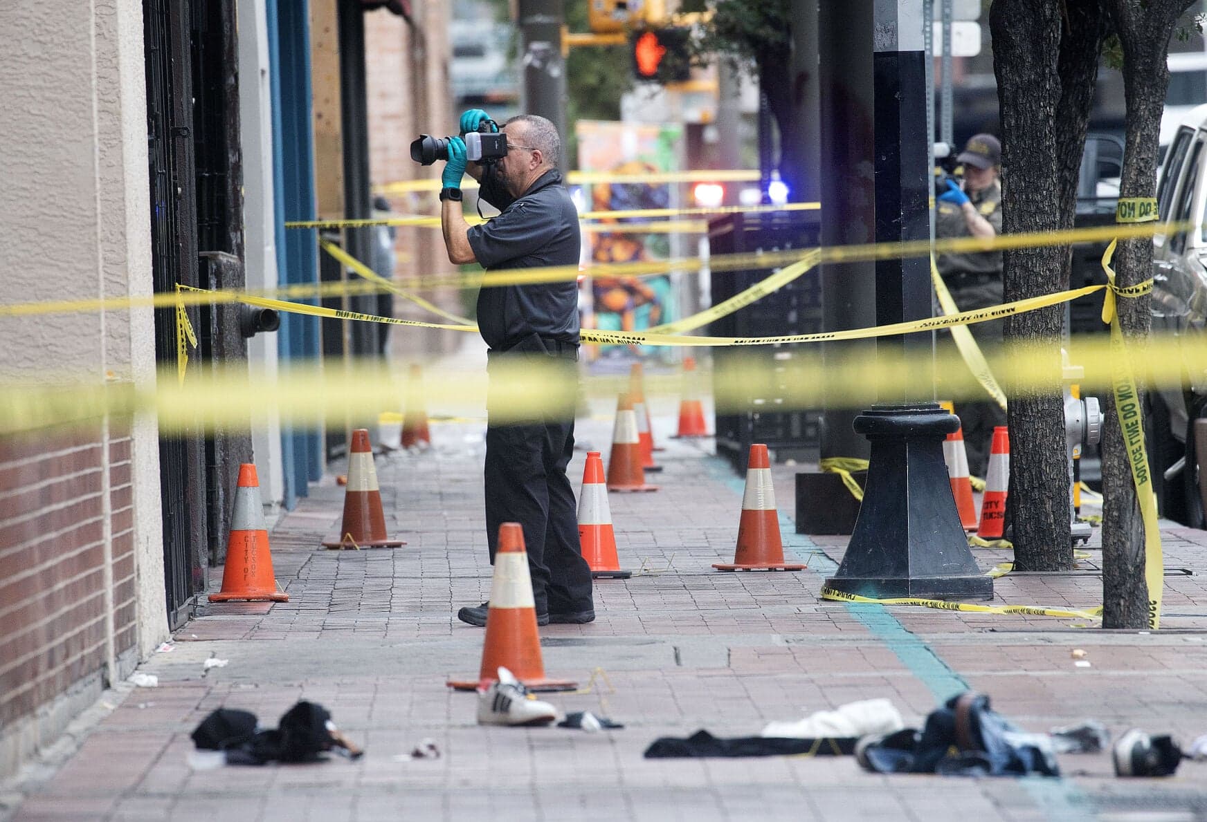 A crime-scene photographer documents evidence on a downtown Tucson sidewalk as police tape and abandoned belongings mark the scene after a shooting that left multiple people critically injured.