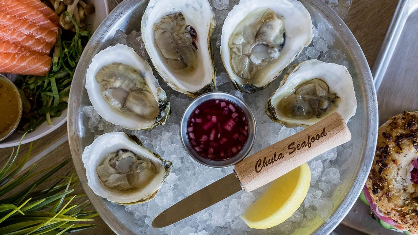 A platter of raw oysters on ice with a Chula Seafood–branded shucking knife, highlighting seafood menu offerings at the chain’s new Arizona Center location.