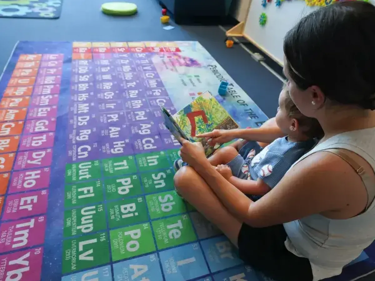 A parent reads with a young child on a colorful periodic-table rug during a Phoenix library summer program encouraging early reading.