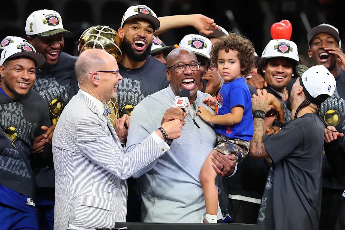 On-court championship celebration after the NBA Finals, with team members surrounding a smiling man holding a child while an interviewer holds a microphone.