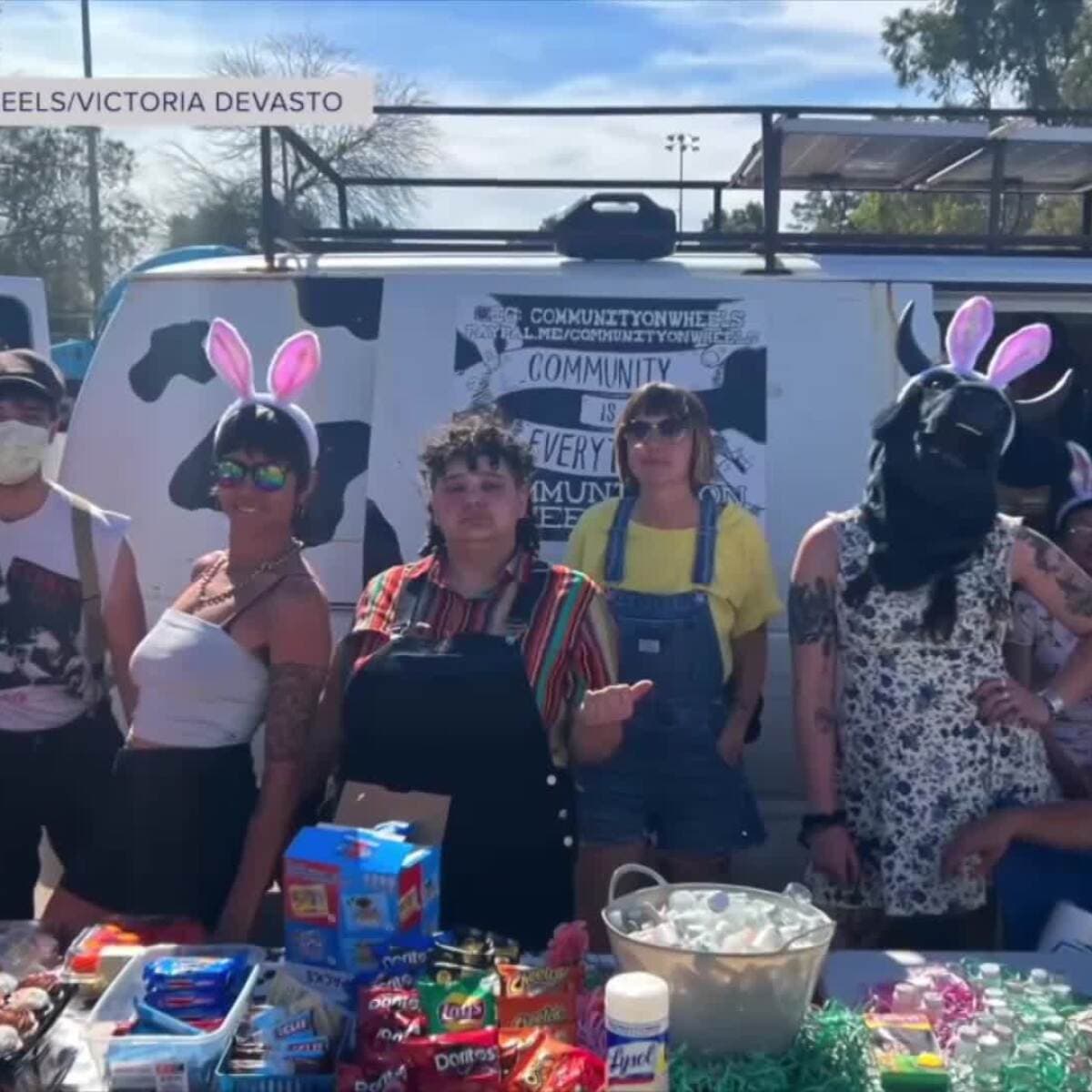 Volunteers from Tucson mutual aid group Community on Wheels stand in front of their cow‑painted van and a table of snacks and supplies at a recent outreach event; the group announced it is shutting down after six years.