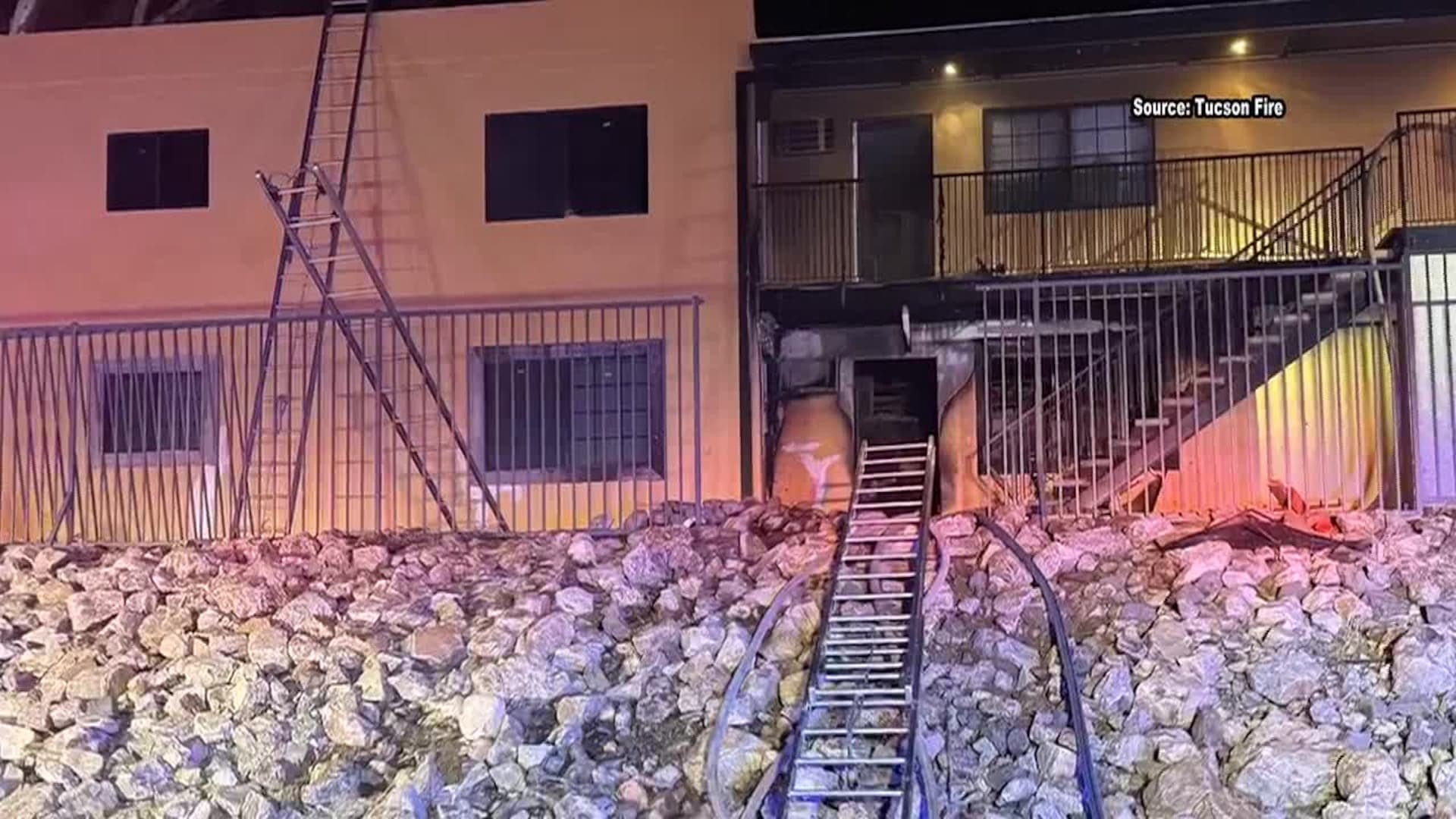 Fire-damaged exterior of a Tucson apartment building showing a charred lower-level unit, a ladder and hoses used by firefighters at the scene.
