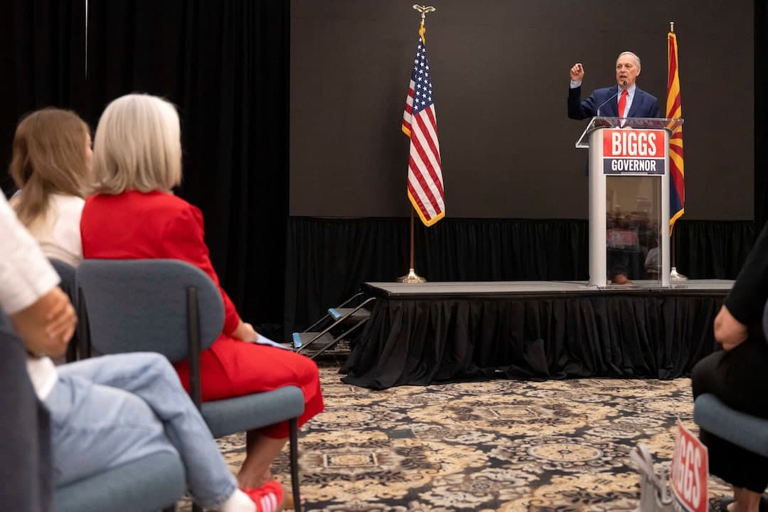 A speaker stands at a podium with a 'Biggs Governor' sign and U.S./Arizona flags at a Republican campaign event in the swing state.