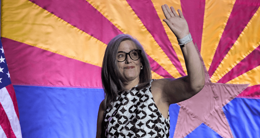 A woman waves onstage in front of an Arizona state flag at a campaign event — image suitable to illustrate coverage of a pay‑to‑play corruption probe involving Arizona’s governor during the election.