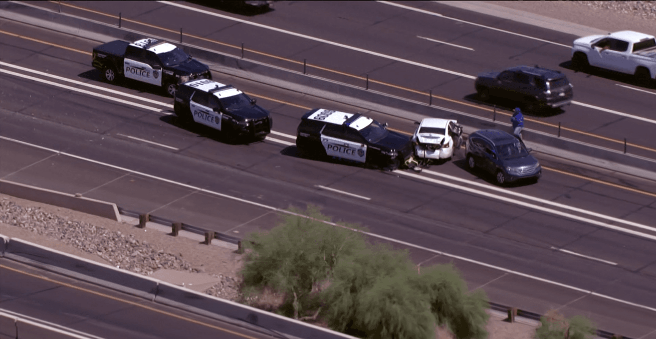 Aerial view showing Mesa Police SUVs and several damaged vehicles blocking multiple lanes on westbound Loop 202 in central Phoenix after a pursuit ended in a multi‑vehicle crash.