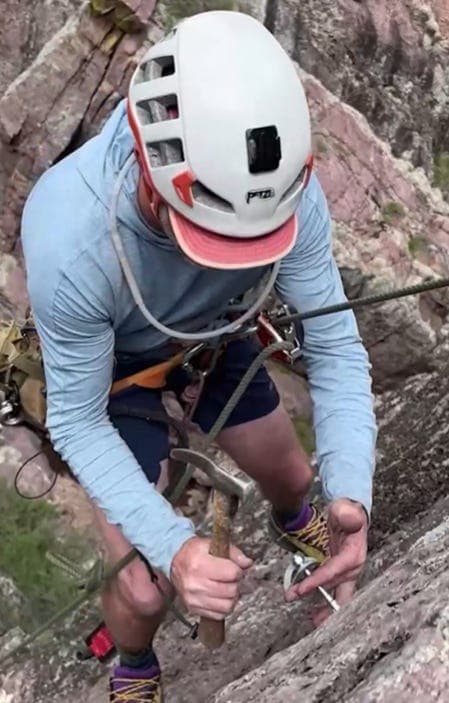 Shows: A route‑setter hammers a bolt into the rock while suspended on a canyon wall, illustrating the hands‑on process of creating new climbing routes.