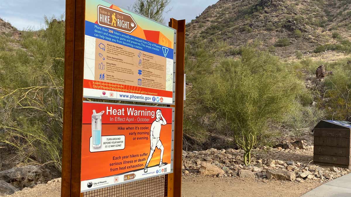 City of Phoenix trailhead sign with a prominent 'Heat Warning' poster advising hikers to avoid midday heat — illustrating local precautions as temperatures soar.