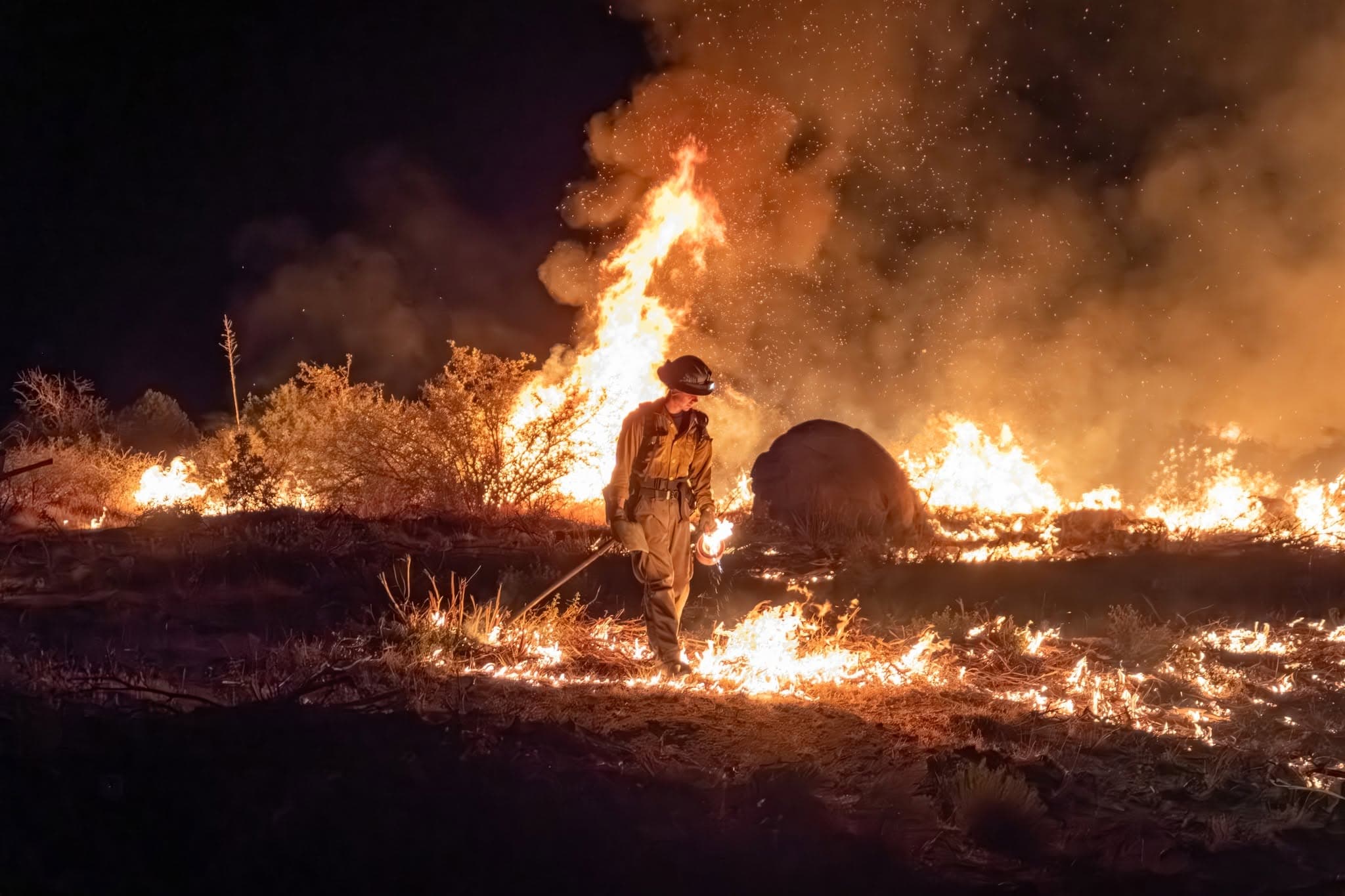 A firefighter walks ahead of a brush fire at night as flames burn nearby — Phoenix fire crews say extreme temperatures increase dangers, especially when combined with substance use.