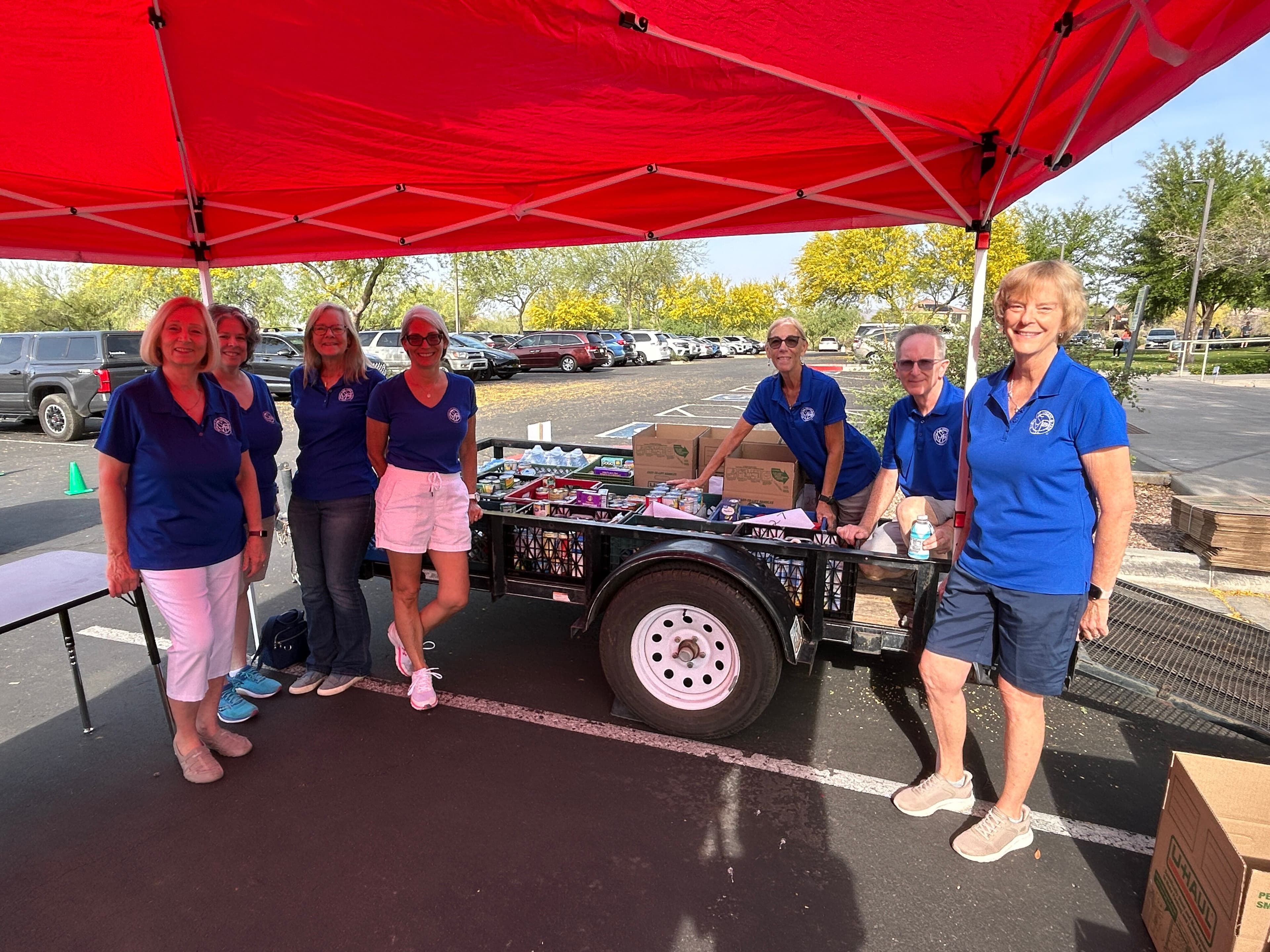 Volunteers in matching shirts stand behind a trailer loaded with canned goods and boxed donations at the event collection point.
