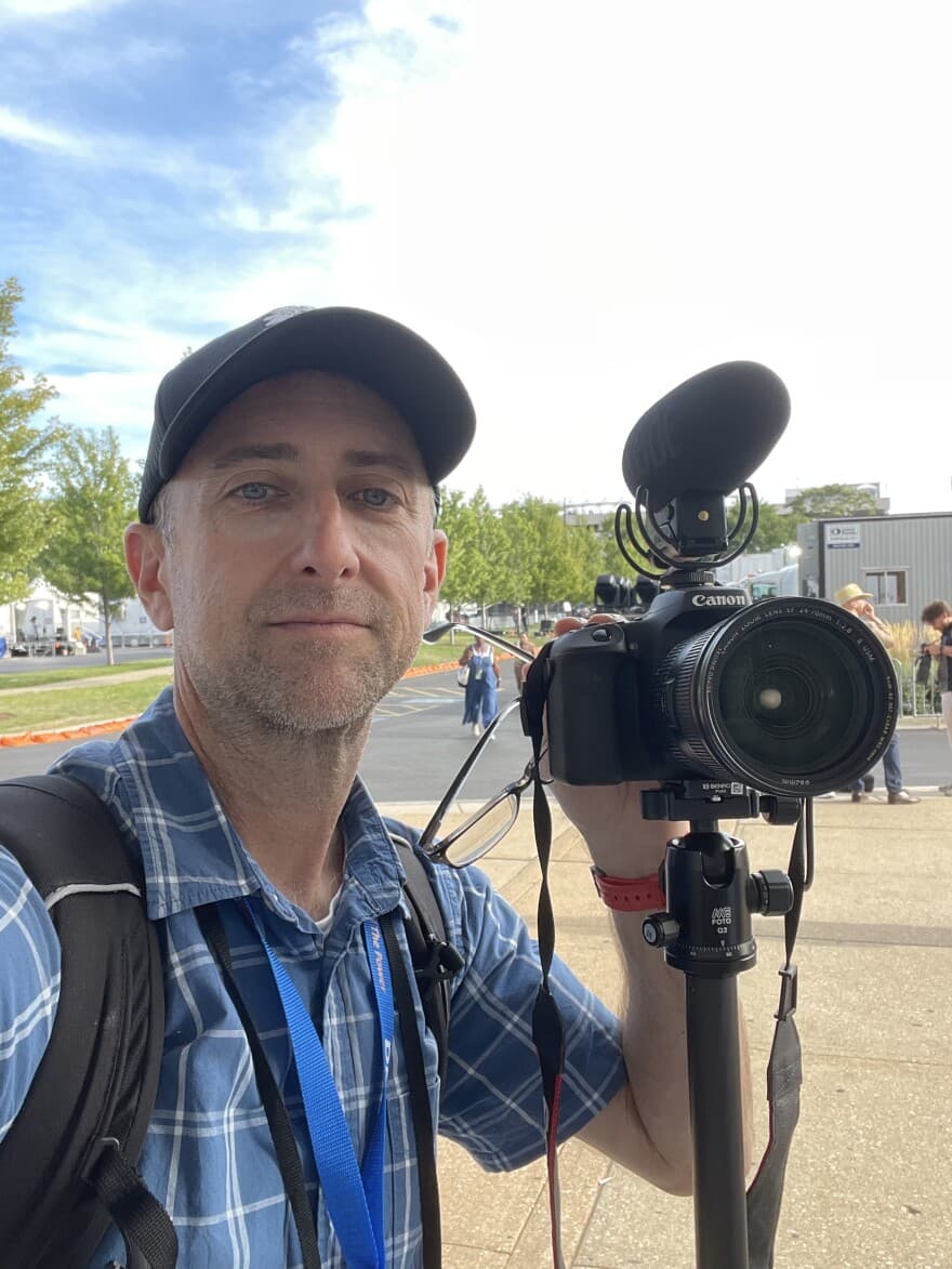 A man holding a video camera with mounted microphone outside an event — the Tucson high school teacher who travels to political conventions to report back to his students.