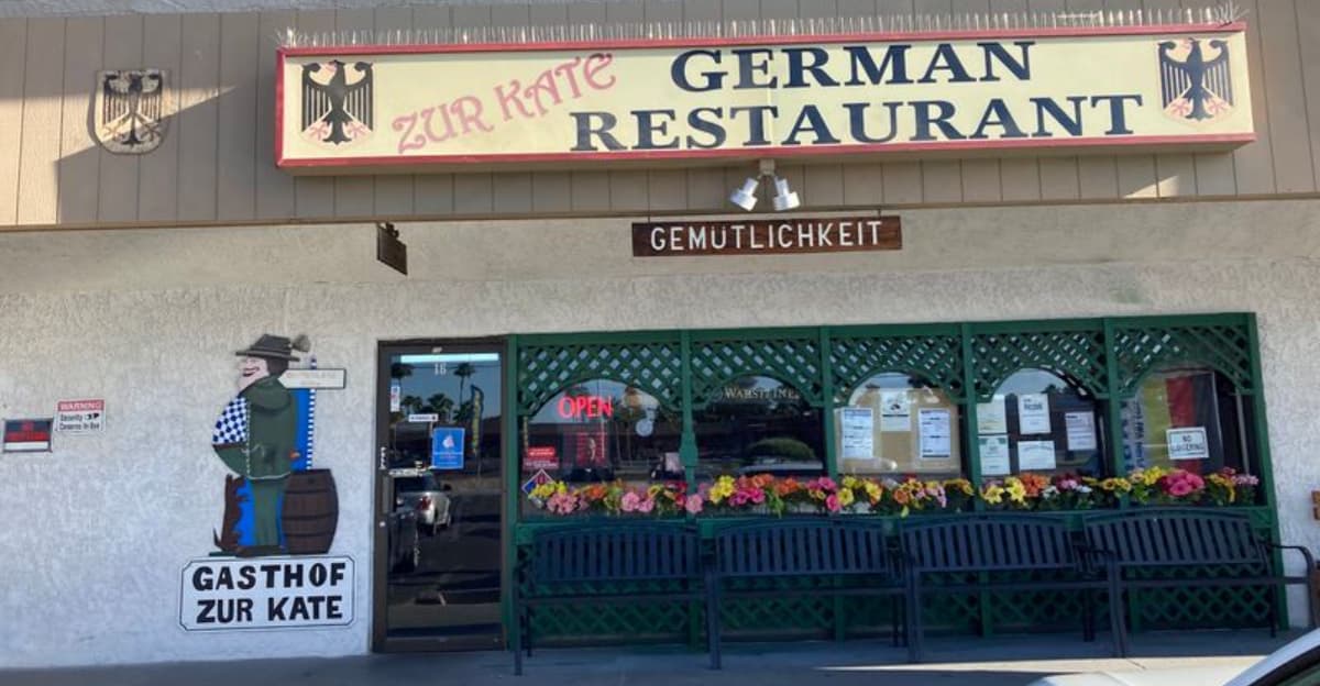 Exterior of Gasthof Zur Kate in Arizona — the German restaurant featured for its Bavarian-style schnitzel, showing the storefront signage, flower boxes and an 'OPEN' neon sign.