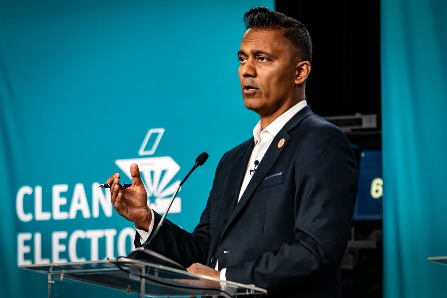 Democratic candidate speaks at a podium during a campaign event, gesturing in front of a ‘Clean Elections’ backdrop.