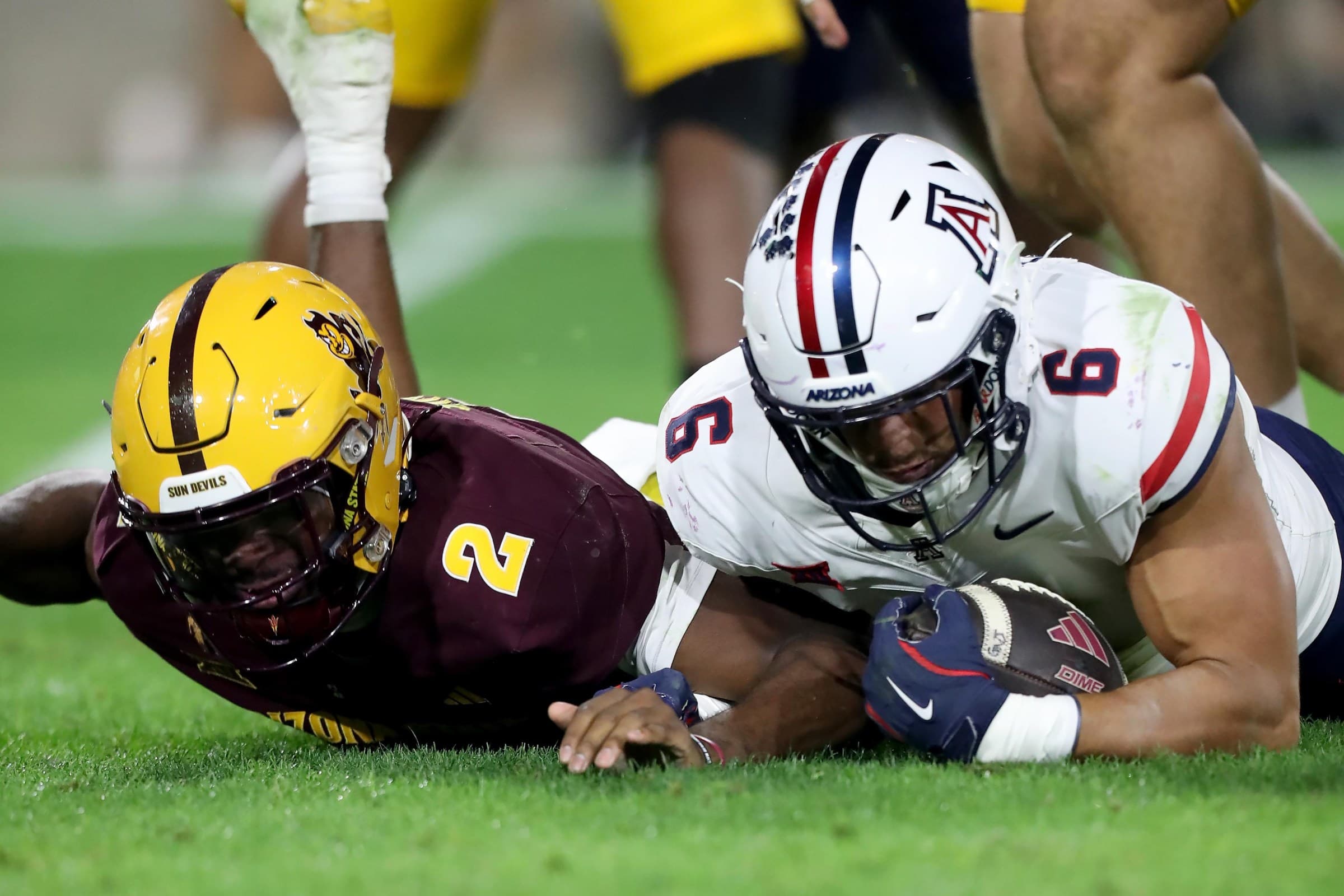 Arizona ballcarrier (white helmet) fights for extra yards while pinned to the turf against an Arizona State defender — a physical run that illustrates the kind of short-yardage and contact work the Wildcats' running back room will be counted on for entering 2026 training camp.