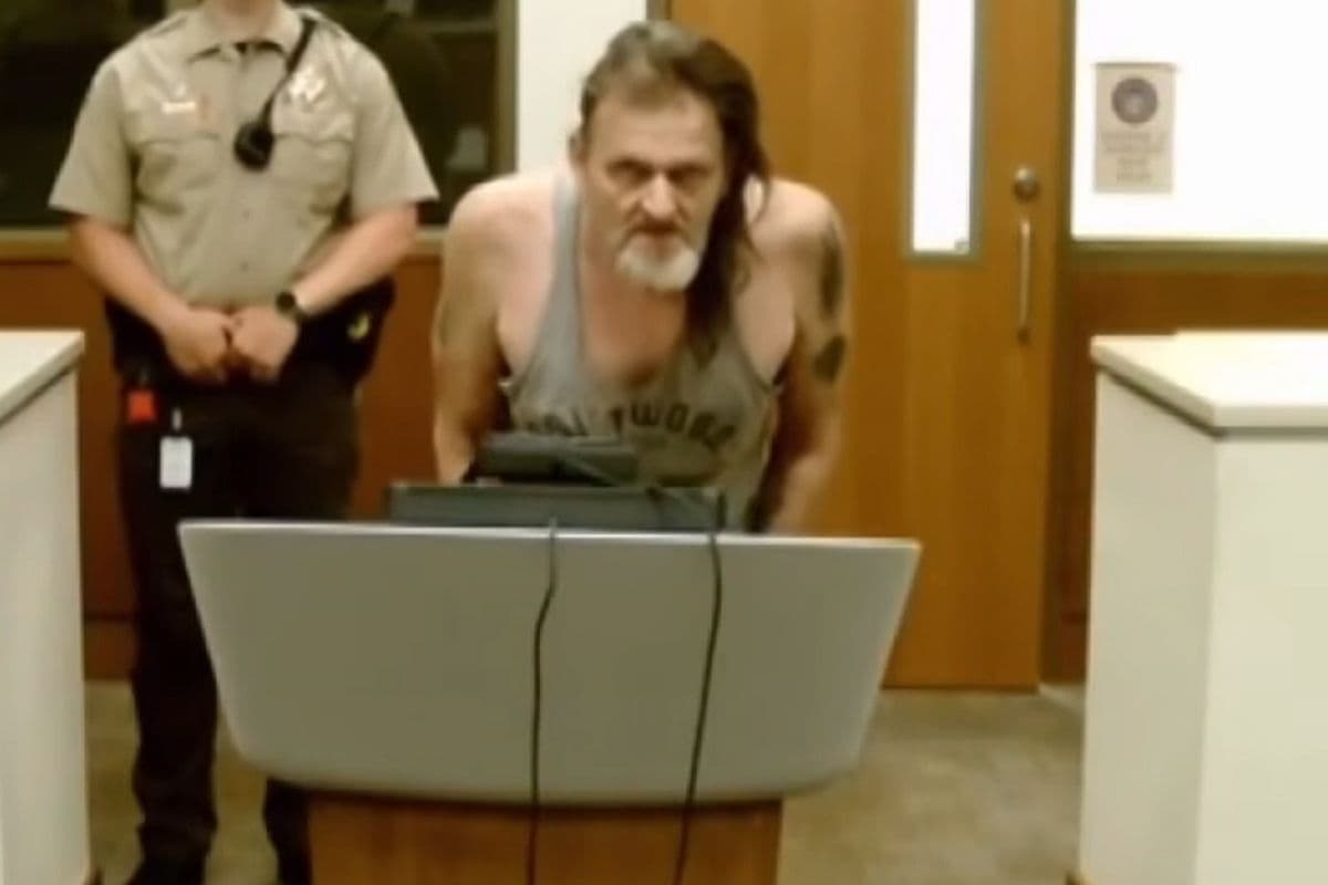 The accused man appears at a court hearing, standing before a podium with an officer behind him; he faces a vulnerable-adult abuse charge over the alleged neglect of his son.