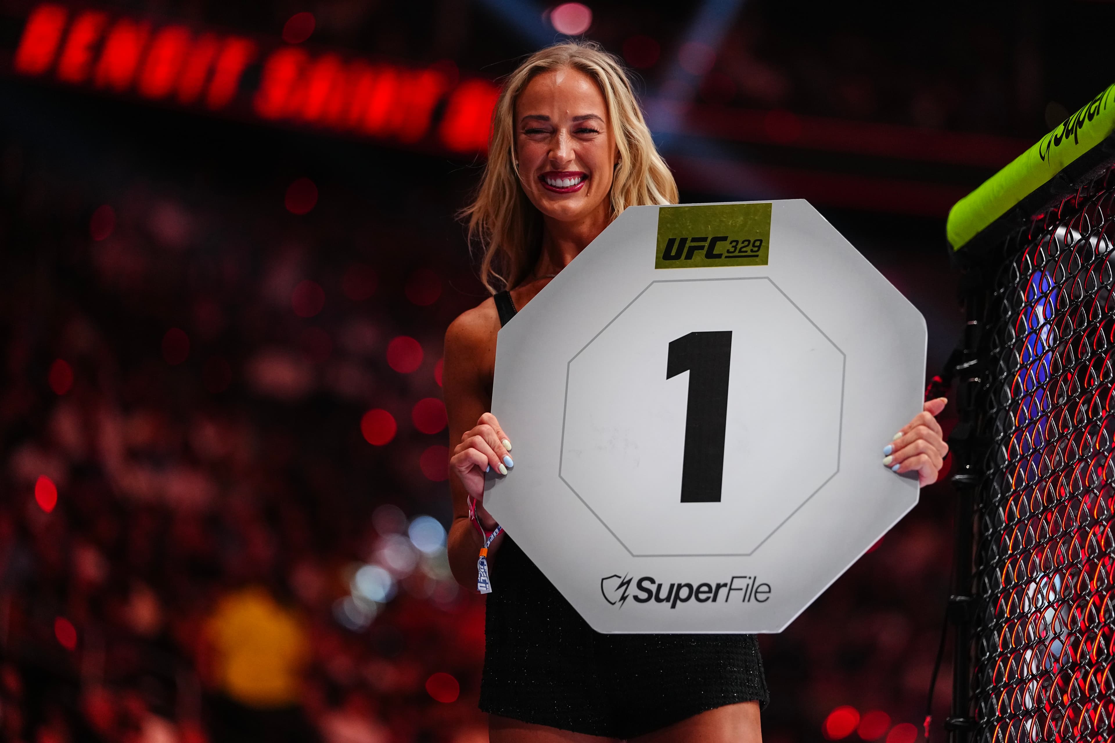 Ring-card holder raises the '1' round sign with UFC329 branding inside the octagon at a live event.