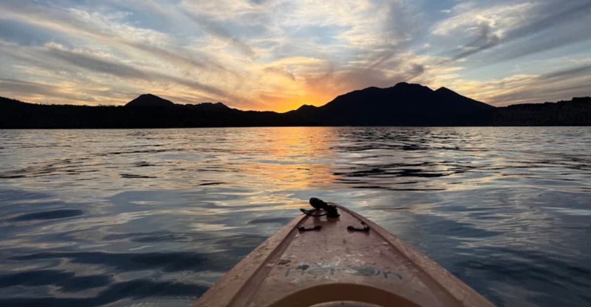 A kayak bow on calm water at dusk with silhouetted mountains — a scene representative of Arizona’s popular evening/glowing night kayak excursions.