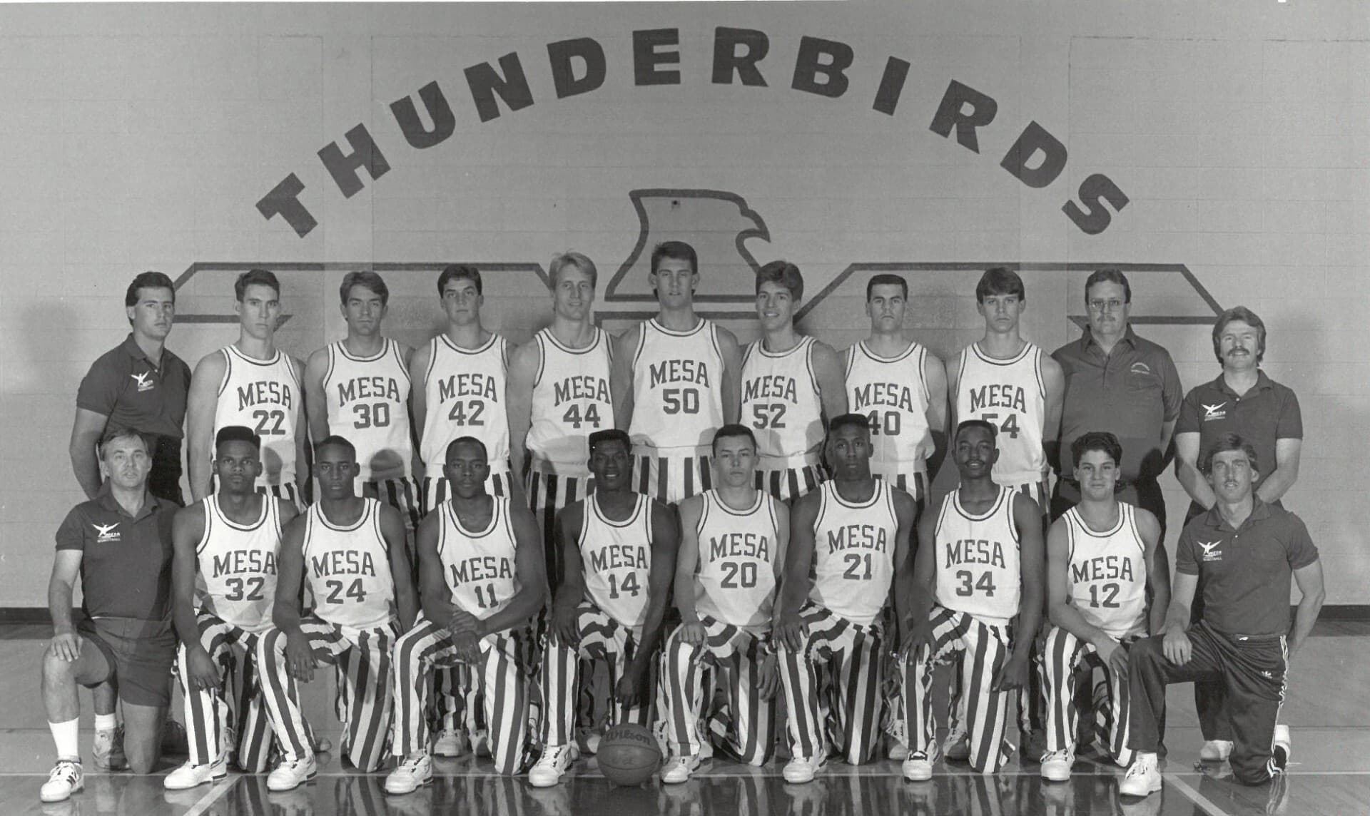 Mesa Community College men's basketball team portrait from the early 1990s, players and coaches posed beneath a 'Thunderbirds' mural in the school gym.