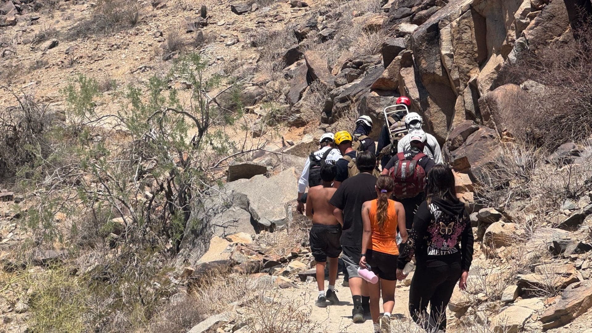 Phoenix rescuers carry a hiker on a litter along a rocky South Mountain trail as bystanders follow; the hiker was later transported to a local hospital in stable condition.