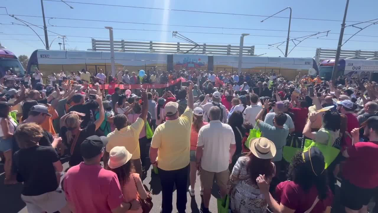 Residents and officials pack the platform for a ribbon-cutting and anniversary event at the South Phoenix light rail extension, with light-rail vehicles visible in the background.