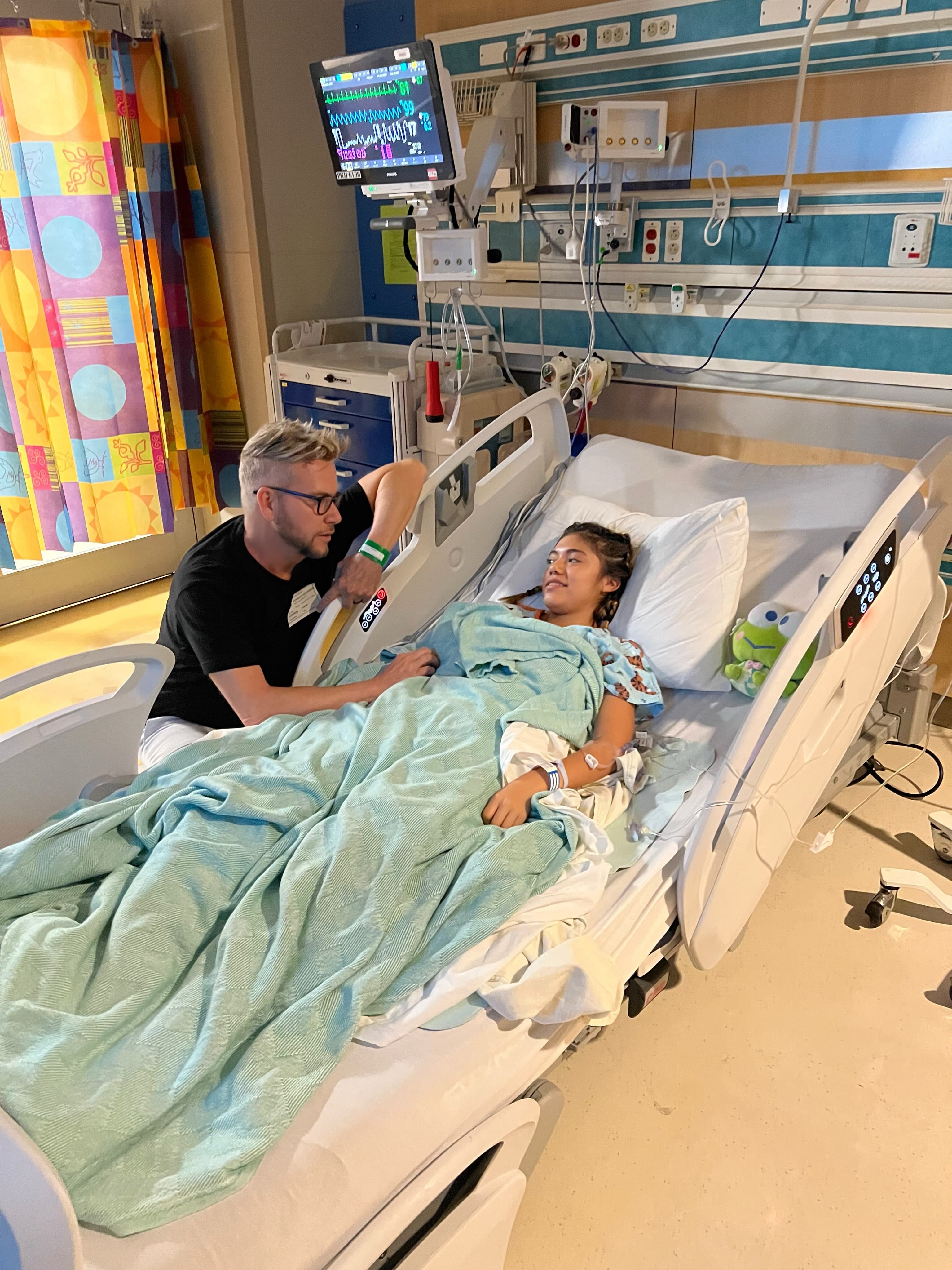 A family member leans at the bedside of a teenage patient in a Phoenix-area hospital room, with monitors and IV lines visible as loved ones gather after securing emergency entry to be reunited.