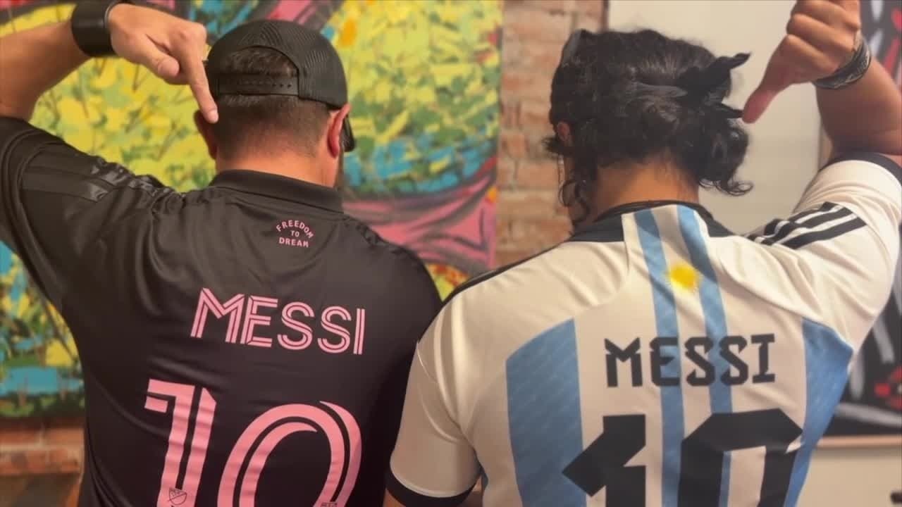 Two World Cup supporters wearing Lionel Messi jerseys pose with their backs to the camera inside Lost Barrio Gallery during a community watch party.