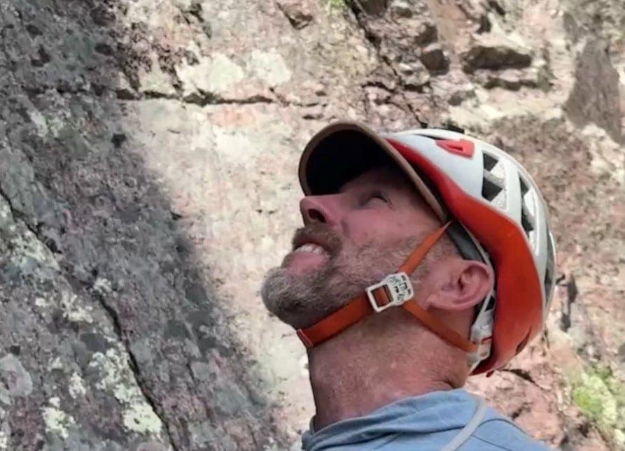 Shows: A route-setter in a helmet looks up at a canyon face while preparing to install climbing hardware during route‑setting work in Arizona.